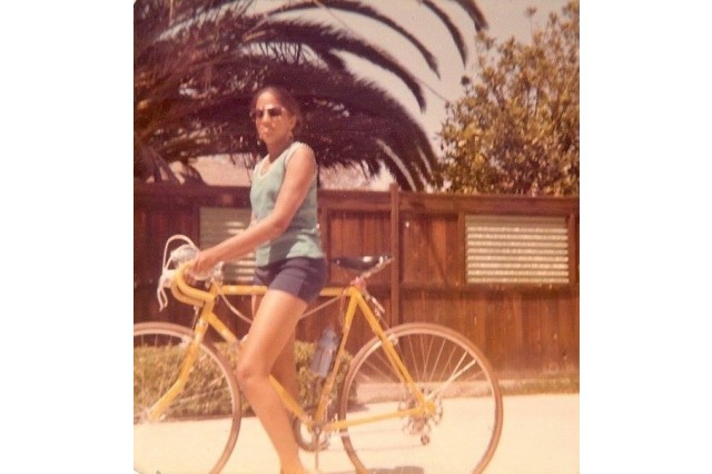 Young woman of color posing on a yellow road bike in front of a wooden fence and palm trees. She wears sunglasses, a green tank top and black shorts.