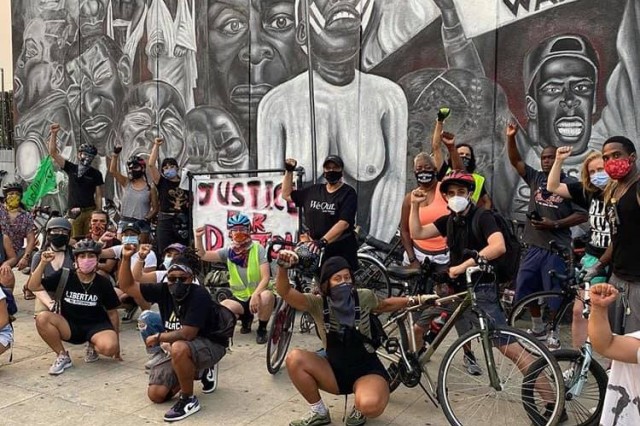 A large group of bike riders, some kneeling and some standing, with fists raised in support of Black Lives Matter. Behind the group is a black and white civil rights mural that says “Freedom Won’t Wait”.