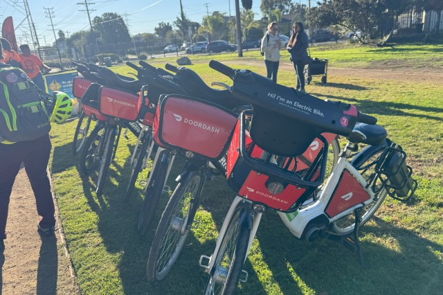 Six white electric bikes with red baskets that have the &quot;Doordash&quot; logo on them parked at an angle on the grass.