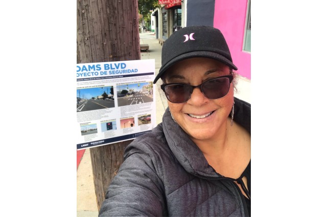 Selfie of a woman of color in a black jacket, baseball cap and sunglasses, standing in front of a bilingual sign for bike safety tacked onto a telephone pole.