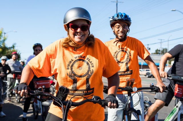 BIPOC mother and son on bikes, standing next to each other. Their shirts say &quot;Biking While Black&quot;