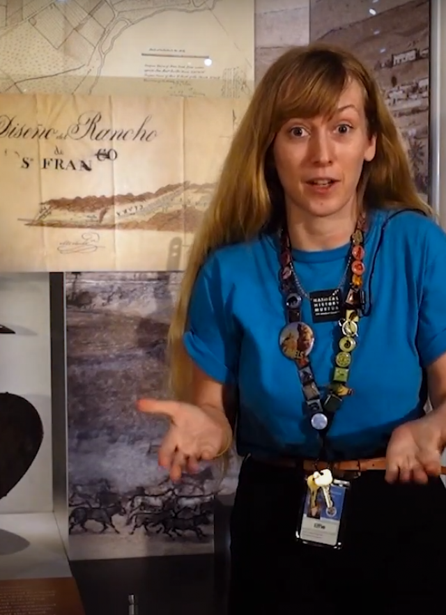 educator stands in front of exhibit case with gold pan and pick axe