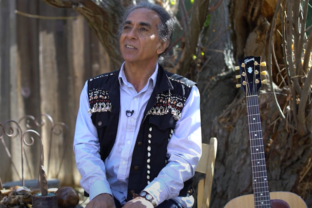 Man with white shirt and black vest. Guitar at side.