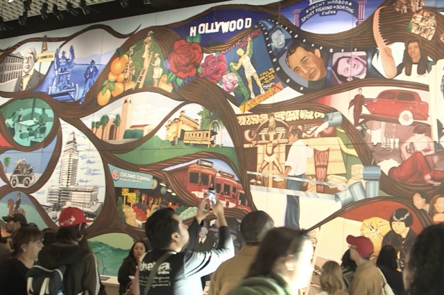 Visitors from the shoulders up viewing a colorful mural made up of tear-shaped vignettes