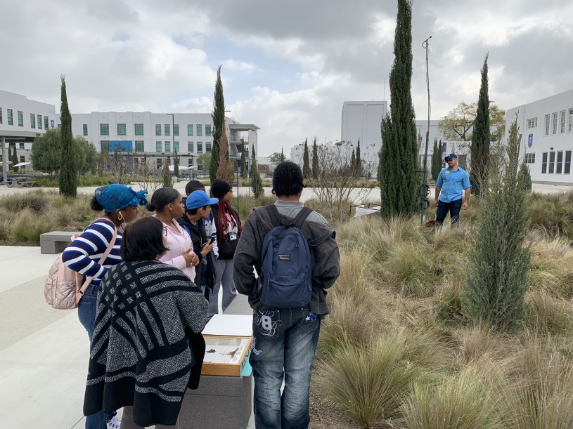 Miguel Ordeñana stands in a green space next to a bat detector while talking to a group of students