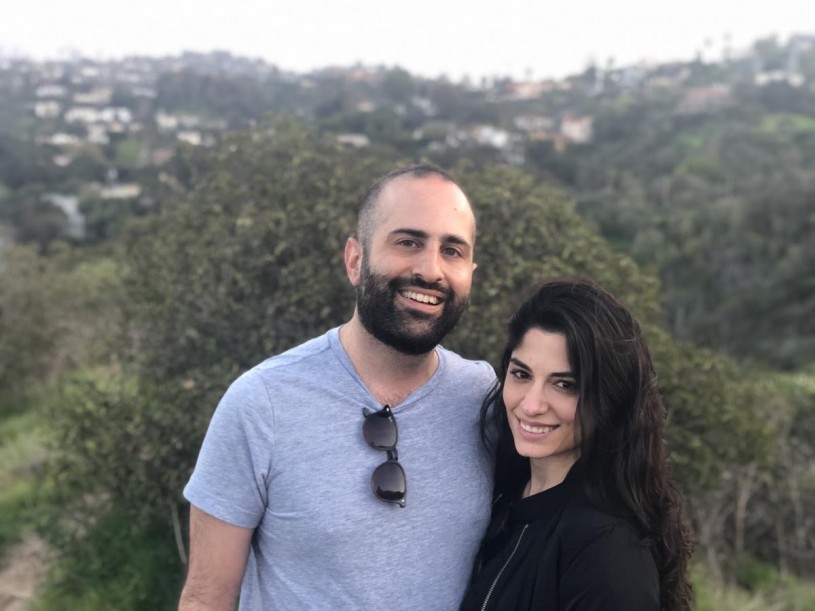 A smiling couple facing the camera stand in nature with a view of the city in the background